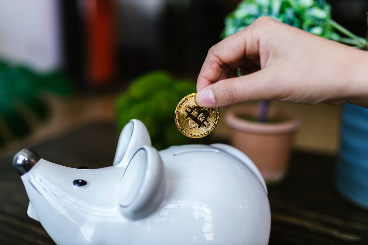 A hand placing a Bitcoin coin into a white piggy bank symbolizing savings.